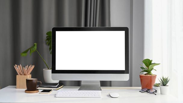 Vista frontal de la computadora simulada con pantalla en blanco, planta de la casa y suministros de oficina en escritorio de oficina blanco. - Foto, Imagen