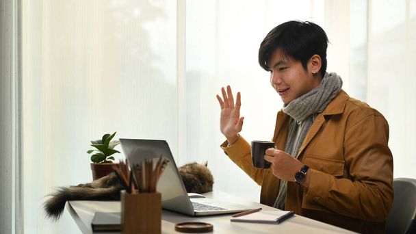Asiatischer Mann telefoniert per Video am Laptop und grüßt seine Kollegen. - Foto, Bild