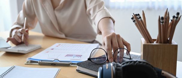 Обрезанный снимок деловой женщины, берущей очки и использующей калькулятор для расчета на рабочем столе. - Фото, изображение