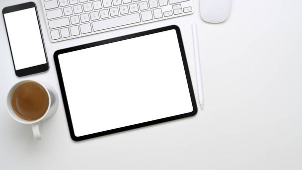 Bovenaanzicht van minimale werkplek met digitale tablet, smartphone, toetsenbord en koffiebeker op wit bureau. - Foto, afbeelding
