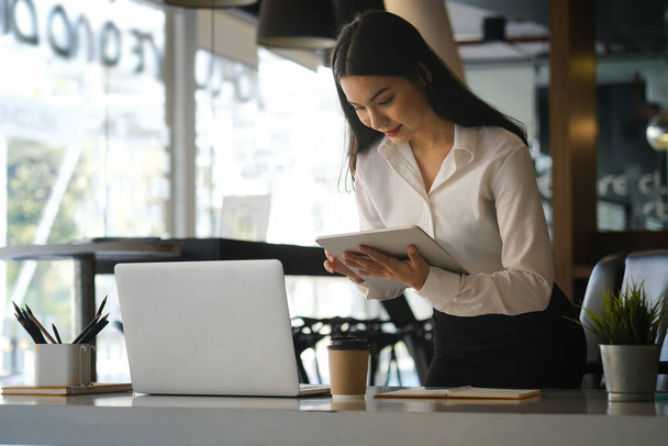 Уверенная молодая деловая женщина, стоящая на рабочем месте и работающая над цифровым планшетом. - Фото, изображение