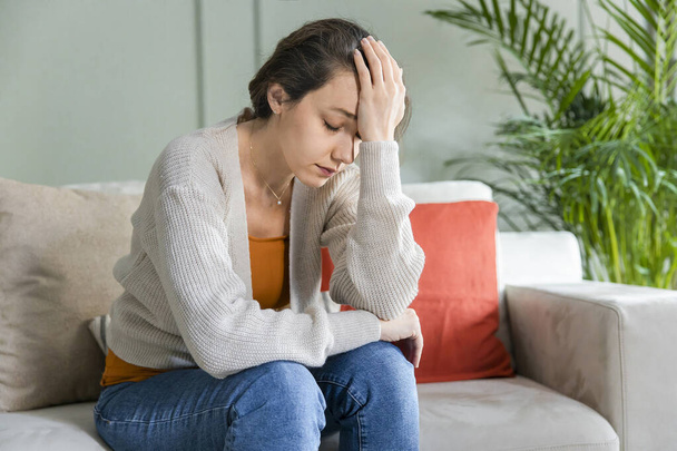 Young depressed woman sitting on sofa in living room. She feeling sad and worried suffering depression in mental health. Problems and broken heart concept. - Foto, imagen
