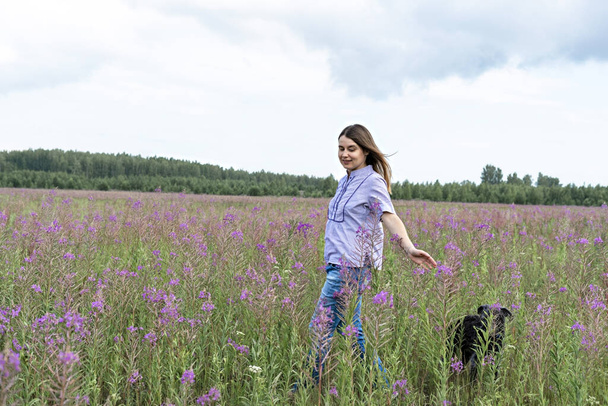 Happy young blond smiling woman in purple shirt walking on a field among flowers of fireweed with a fluffy gray dog, traveling with pets friendship - Photo, Image