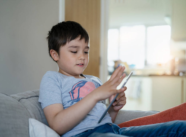 Aito Kid istuu sohvalla katsomassa sarjakuva tai pelaa peliä tabletilla, Lapsipoika käyttää digitaalista pad oppimisen oppitunti verkossa internetissä, Kotikoulutus, Etäopetus concep - Valokuva, kuva