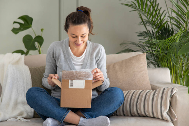 Young woman in her living room looks happy as she unpacks a small cardboard box containing something exciting - Photo, Image