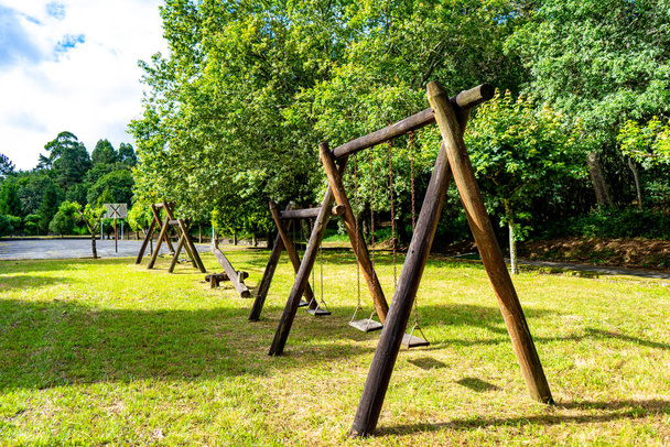 solitario parque infantil con columpios de madera en un lugar muy verde y con un campo de delporte al fondo - Fotografie, Obrázek