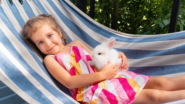 Tyttö, jolla on valkoinen pupu, makaa riippumatossa kesällä aurinkoisena päivänä. Valkoinen pörröinen koristeellinen kani pienen tytön sylissä. Jänis kuin lemmikki. Lapsi pitelee pientä jänistään käsissään.. - Valokuva, kuva