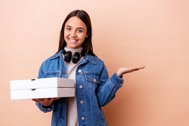 Joven mujer de raza mixta sosteniendo una pizza aislada mostrando un espacio de copia en una palma y sosteniendo otra mano en la cintura. - Foto, imagen