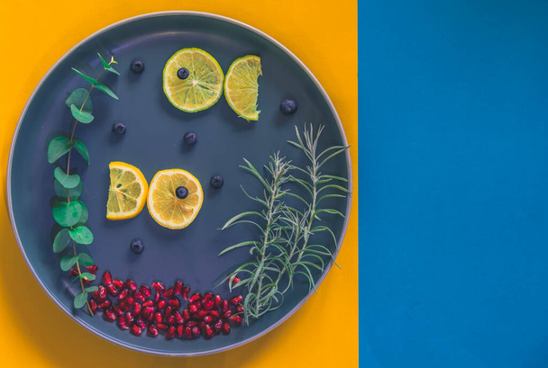 citromszeletekből származó halak, amelyek zöld növényekkel úszkálnak a tányéron, a kék és sárga háttérrel elszigetelt terület másolása - Fotó, kép