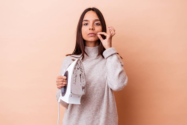 Young mixed race woman holding an iron isolated with fingers on lips keeping a secret. - Photo, Image