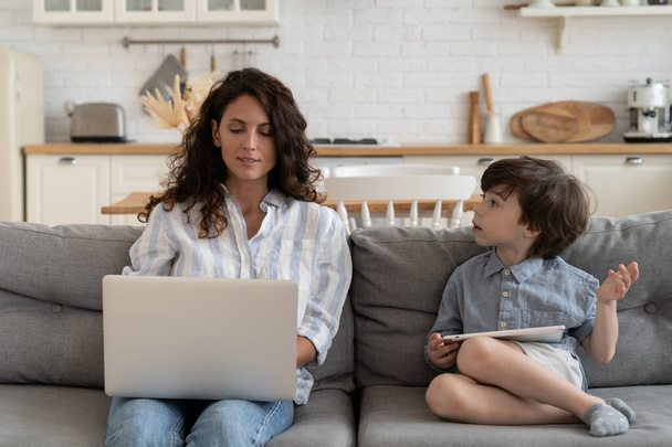 A kissrác anyával beszél, aki laptopon gépel. Multitasking anya üzletasszony dolgozik otthonról a fiával - Fotó, kép