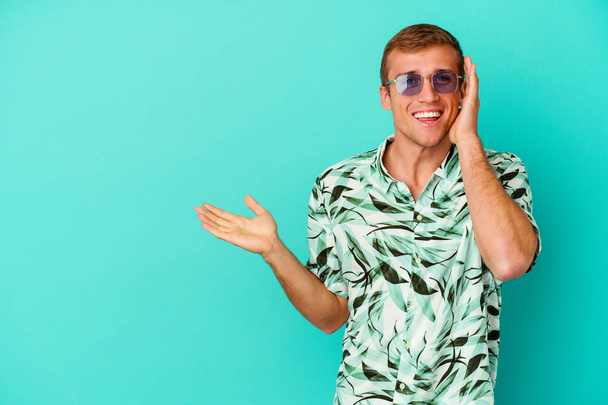 Young caucasian man wearing a summer clothes isolated on blue background holds copy space on a palm, keep hand over cheek. Amazed and delighted. - Photo, image