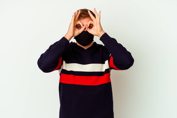 Junger kaukasischer Mann mit Maske für Virus isoliert auf weißem Hintergrund, der Okay-Zeichen über den Augen zeigt - Foto, Bild