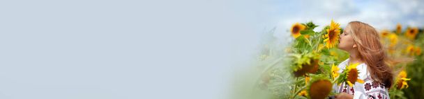 Una hermosa chica en una camisa bordada con el pelo revoloteando huele una flor de girasol. Día de la Independencia de Ucrania. Postal, cartel, calendario - Foto, imagen