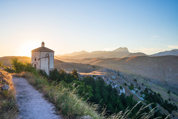 Το μικρό οκταγωνικό εκκλησάκι κοντά στο κάστρο Rocca Calascio ερείπια κατά το ηλιοβασίλεμα στο παρασκήνιο, ορόσημο στο Εθνικό Πάρκο Gran Sasso, Abruzzo, Ιταλία. Γραφικό φόντο βουνών. - Φωτογραφία, εικόνα