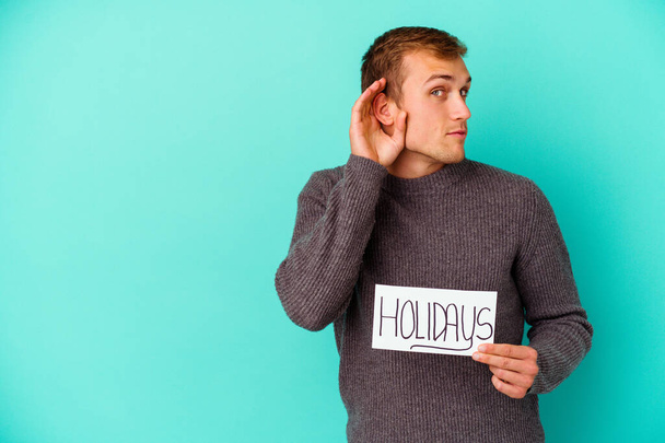 Jovem caucasiano segurando um cartaz Holidays isolado no fundo azul tentando ouvir uma fofoca. - Foto, Imagem