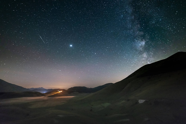 Πανοραμική πρόσκρουση νυχτερινού ουρανού και μετεωριτών πάνω από τα υψίπεδα Castelluccio di Norcia, Ιταλία. Ο γαλαξίας Milky Way τόξο και τα αστέρια πάνω από φωτισμένο χωριό μοναδικό τοπίο λόφους. Δίας πλανήτης ορατός. - Φωτογραφία, εικόνα