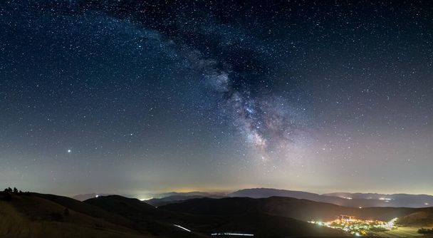 イタリアのSanto Stefano di Sessanio 、 Abruzzo 、 Rocca Calascioのパノラマの夜空。天の川銀河弧と照明村のユニークな丘の風景の上の星。木星の惑星が見える. - 写真・画像