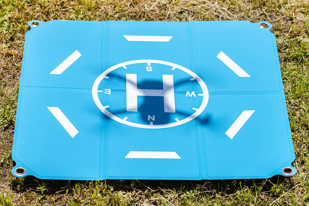 Cień startującego drona jest widoczny na niebieskim lądowisku leżącym na trawie.. - Zdjęcie, obraz