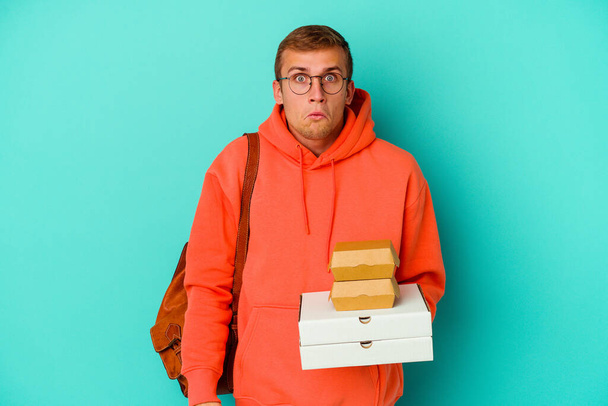 Young student caucasian man holding hamburgers and pizzas isolated on blue background shrugs shoulders and open eyes confused. - Photo, Image