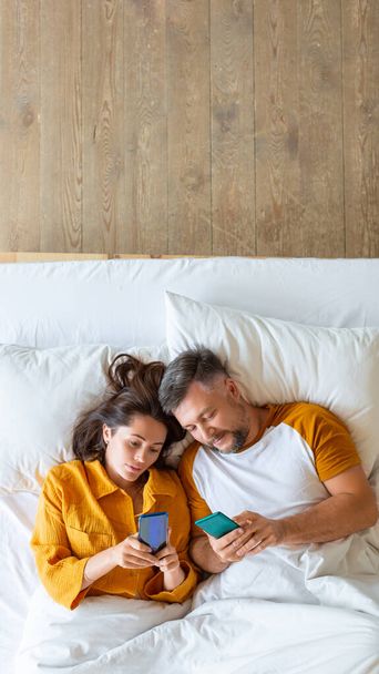 Foto verticale di una coppia di mezza età sdraiata a letto con smartphone. - Foto, immagini