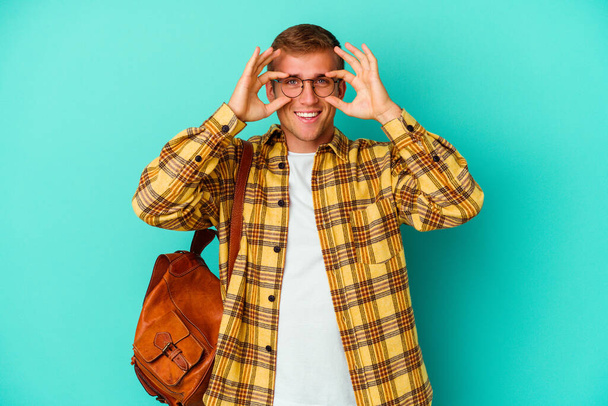 Young caucasian student man isolated on blue background showing okay sign over eyes - Φωτογραφία, εικόνα