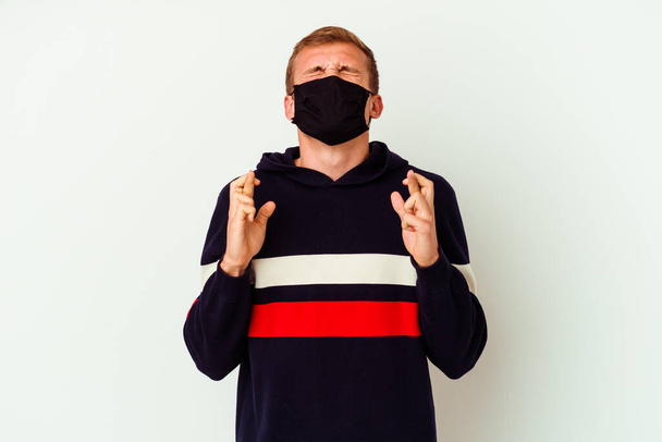 Молодий білий чоловік у масці для вірусу ізольований на білому тлі, що перетинає пальці, щоб мати удачу
 - Фото, зображення