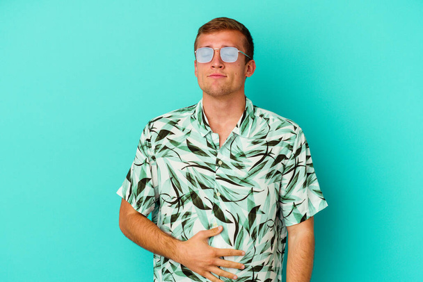 Young caucasian man wearing a summer clothes isolated on blue background touches tummy, smiles gently, eating and satisfaction concept. - Photo, Image