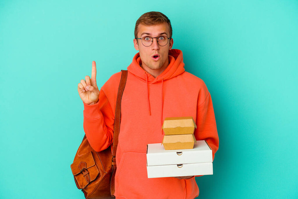 Young student caucasian man holding hamburgers and pizzas isolated on blue background having some great idea, concept of creativity. - Foto, imagen