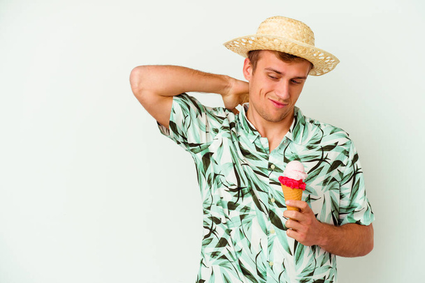 Молодой кавказский мужчина, одетый в летнюю одежду и держащий мороженое на белом фоне, трогает затылок, думая и делая выбор. - Фото, изображение