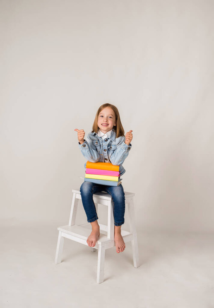 a happy little blonde girl in denim is sitting with textbooks on a white background with a place for text. Back to school - Foto, imagen