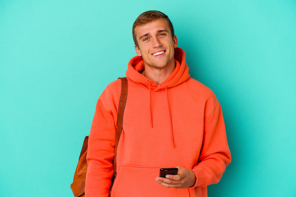 青の背景に孤立した携帯電話を持っている若い学生白人男性幸せ、笑顔と陽気な. - 写真・画像