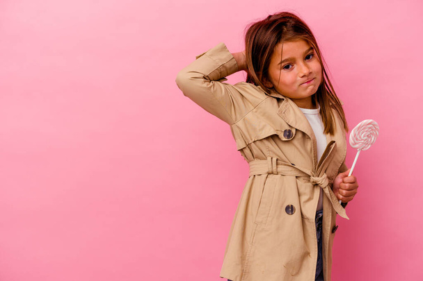 Kleines kaukasisches Mädchen mit einem Lutscher auf rosa Hintergrund, der den Hinterkopf berührt, denkt und eine Wahl trifft. - Foto, Bild