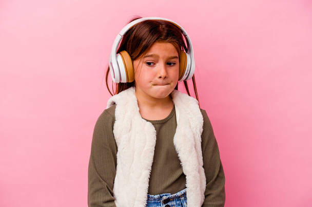 Piccola ragazza caucasica ascoltare musica isolata su sfondo rosa confuso, si sente dubbioso e insicuro. - Foto, immagini