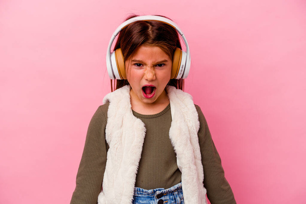 Little caucasian girl listening music isolated on pink background screaming very angry and aggressive. - Fotoğraf, Görsel