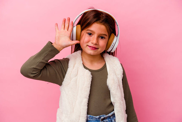 Маленькая кавказская девочка слушает музыку, изолированную на розовом фоне улыбаясь весело показывая номер пять пальцами. - Фото, изображение