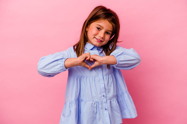 Klein blank meisje geïsoleerd op roze achtergrond glimlachen en het tonen van een hart vorm met handen. - Foto, afbeelding