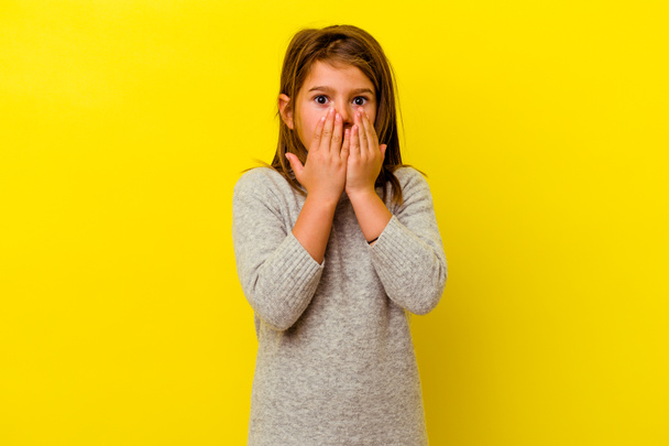 Маленькая кавказская девочка, изолированная на жёлтом фоне, потрясена, закрывая рот руками, желая обнаружить что-то новое.. - Фото, изображение