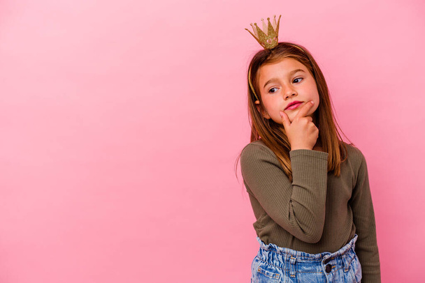 klein prinses meisje met kroon geïsoleerd op roze achtergrond kijken zijwaarts met twijfelachtige en sceptische uitdrukking. - Foto, afbeelding