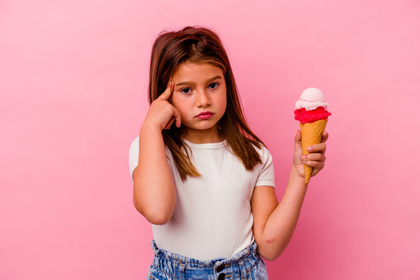 Little caucasian girl holding ice cream isolated on pink background pointing temple with finger, thinking, focused on a task. - Foto, afbeelding