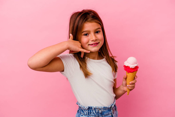Маленькая кавказская девочка держит мороженое на розовом фоне, показывая жест мобильного телефона пальцами. - Фото, изображение