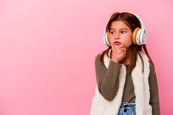 Petite fille caucasienne écoutant de la musique isolée sur fond rose regardant de côté avec une expression douteuse et sceptique. - Photo, image