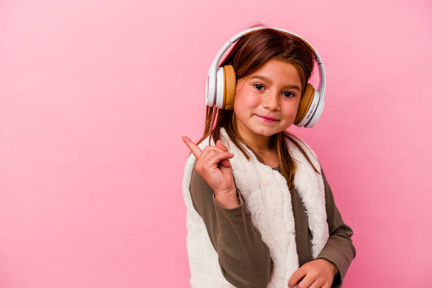 Petite fille caucasienne écoutant de la musique isolée sur fond rose souriant et pointant du doigt, montrant quelque chose à vide. - Photo, image