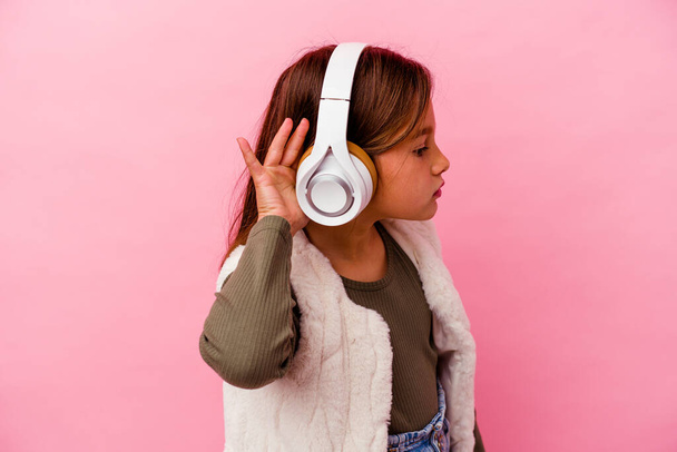 Pequeña chica caucásica escuchando música aislada sobre fondo rosa tratando de escuchar un chisme. - Foto, imagen