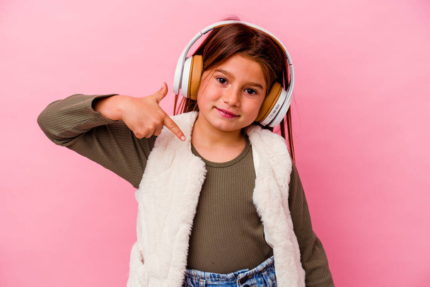Маленькая кавказская девочка слушает музыку, изолированную на розовом фоне, человек указывает рукой на пространство для копирования рубашки, гордый и уверенный - Фото, изображение