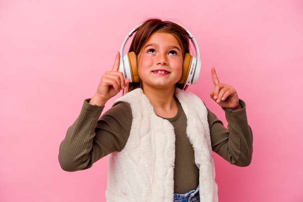 Menina caucasiana ouvindo música isolada no fundo rosa apontando de cabeça com a boca aberta. - Foto, Imagem