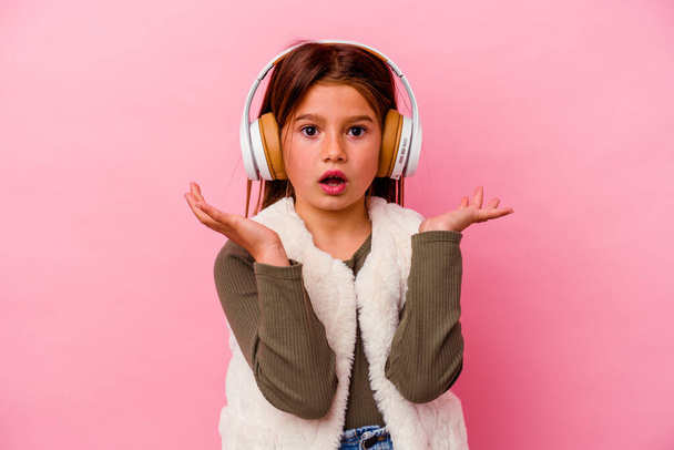 Little caucasian girl listening music isolated on pink background surprised and shocked. - 写真・画像