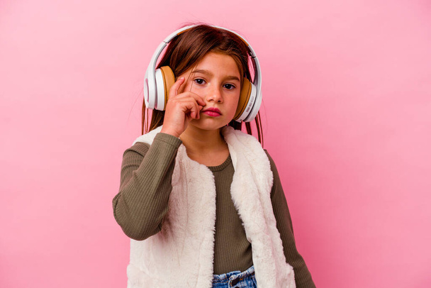 Petite fille caucasienne écoutant de la musique isolée sur fond rose avec les doigts sur les lèvres gardant un secret. - Photo, image