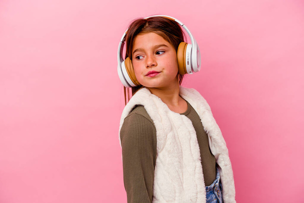 Petite fille caucasienne écoute de la musique isolée sur fond rose regarde de côté souriant, gai et agréable. - Photo, image