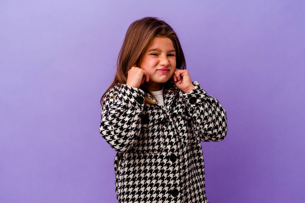 Маленькая кавказская девочка изолирована на фиолетовом фоне Маленькая кавказская девочка изолирована на фиолетовом фоне закрывая уши руками. - Фото, изображение